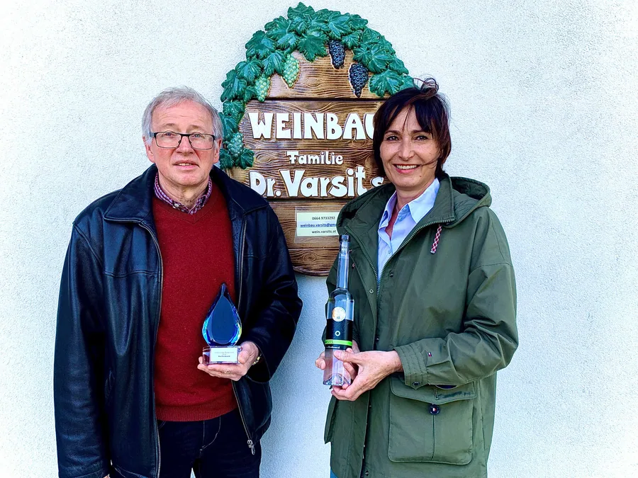 Michael und Martina Varsits vor dem geschnitzten Schild ‚Weinbau Familie Dr. Varsits‘, mit Landessieger-Trophäe und einer Flasche Marillenbrand